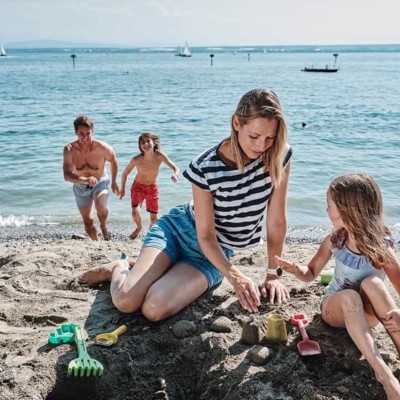 Mutter und Tochter sitzen am Strand, Vater und Sohn kommen aus dem Bodensee gelaufen