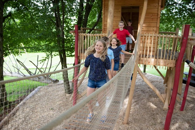 Kinder rennen über eine Hängebrücke im Erlebnis-Baumhaus auf dem Spielplatz. Im Hintergrund liegt ein Baumstamm zum Balancieren.