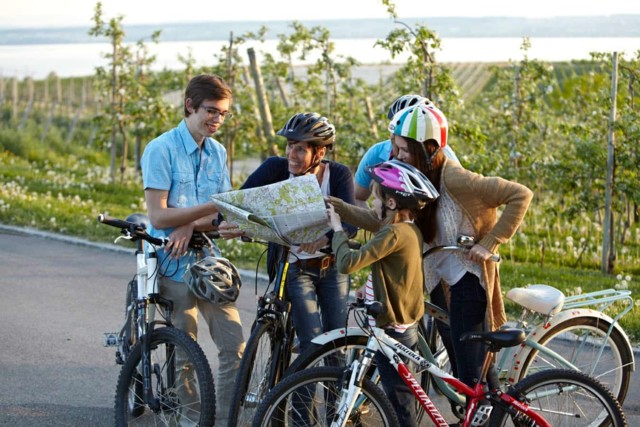 Eine Familie steht in den Obstplantagen am Bodensee und schaut lachend auf eine Radkarte.