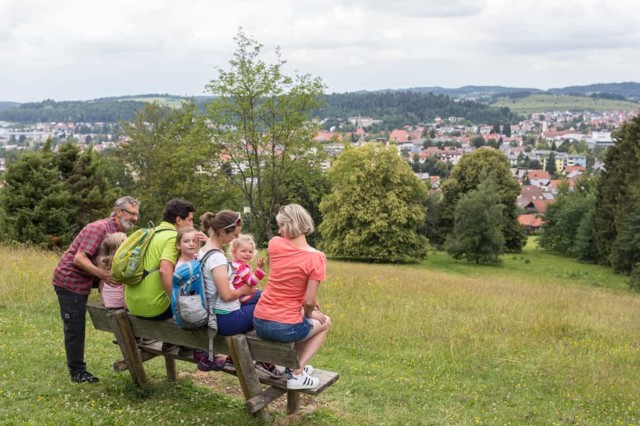 Familien mit Wanderrucksäcken sitzen auf einer Bank mit Blick über Münsingen.