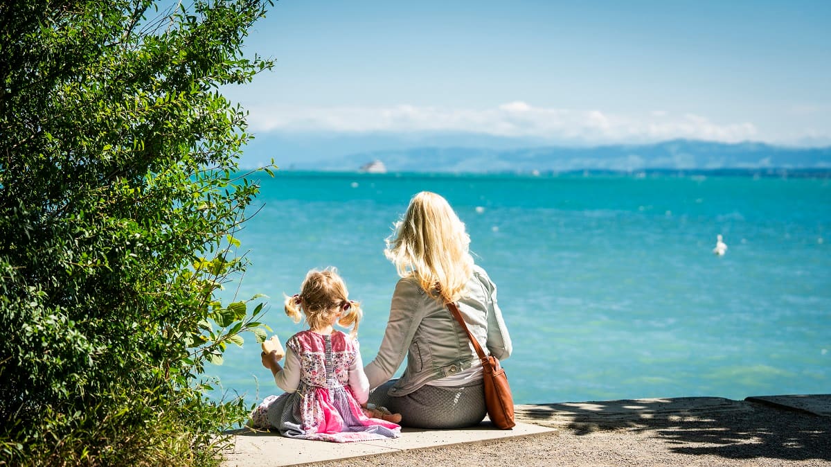 Eine Mutter sitzt  mit ihrer kleinen Tochter am Ufer und blickt auf den Bodensee.