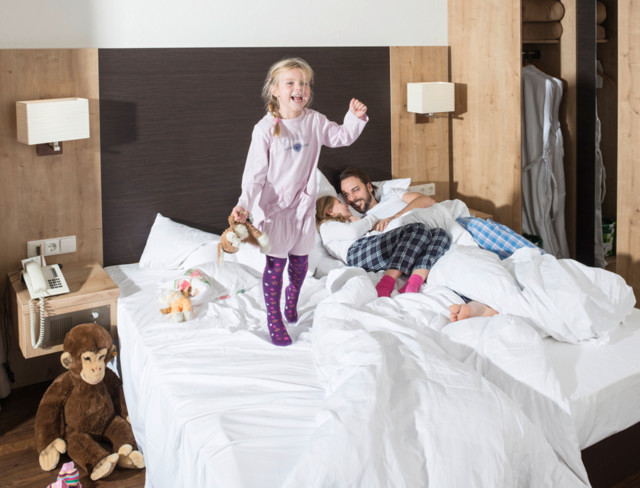 Ein Mädchen mit einem Kuscheltier in der Hand hüpft auf einem Bett. Neben dem Bett sitzt ein Affe.