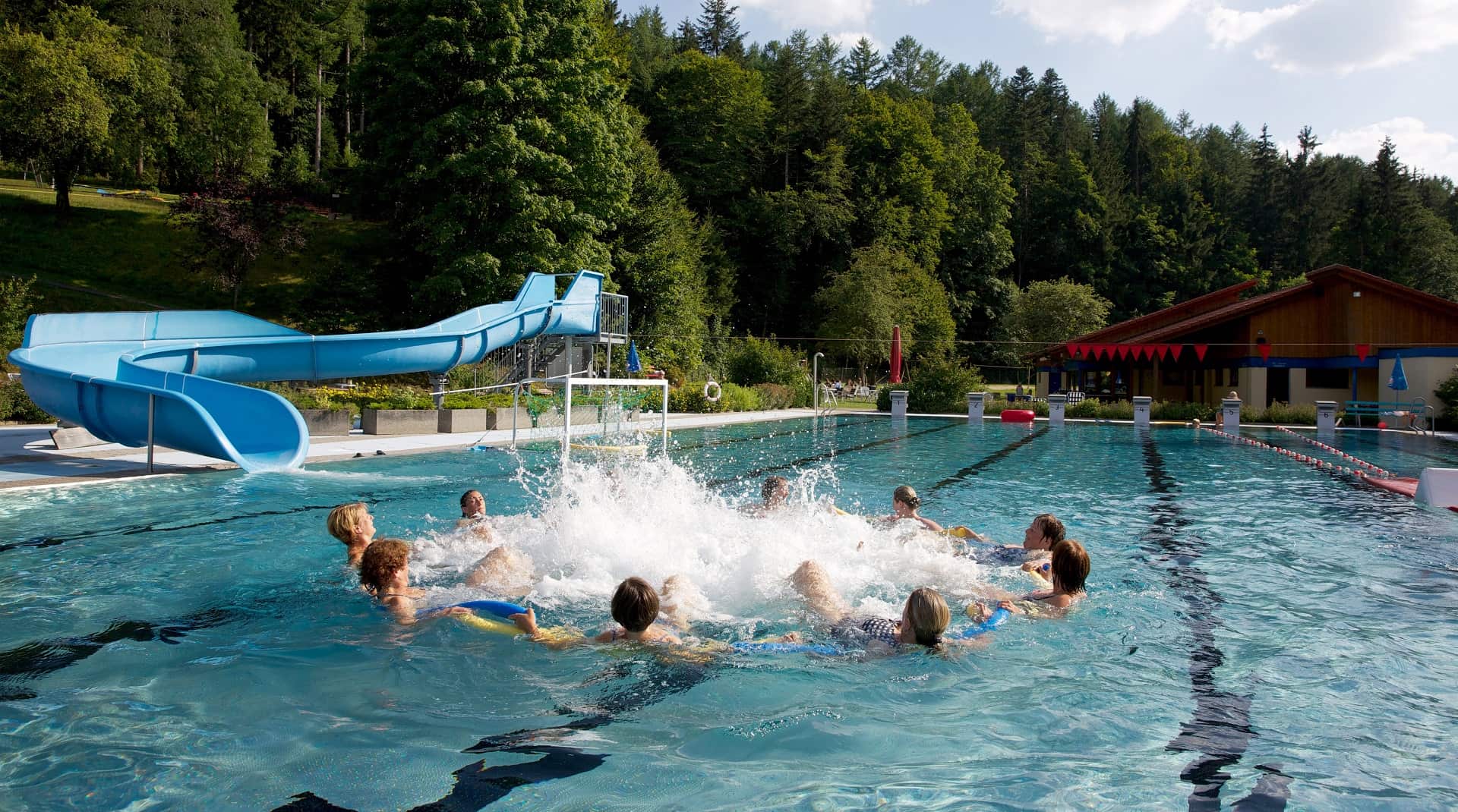 Frauen bilden im Schwimmbecken einen Kreis. Im Hintergrund ist steht die blaue Wasserrutsche ins Freibad Obertal.