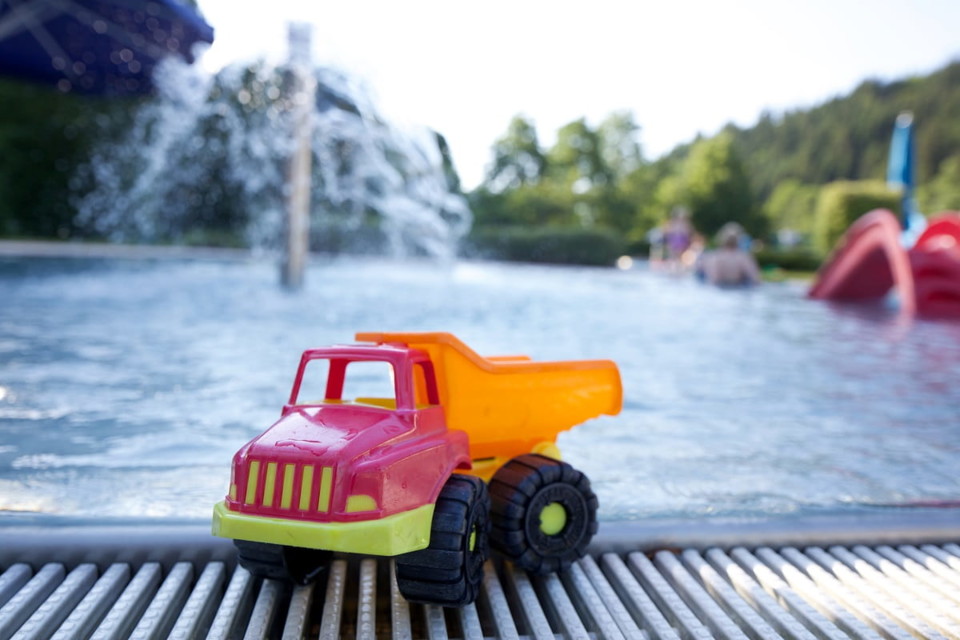 Ein kleiner Spielzeugkipplaster steht am Schwimmbeckenrand. Im Hintergrund steht eine rote Wasserrutsche für kleine Kinder.