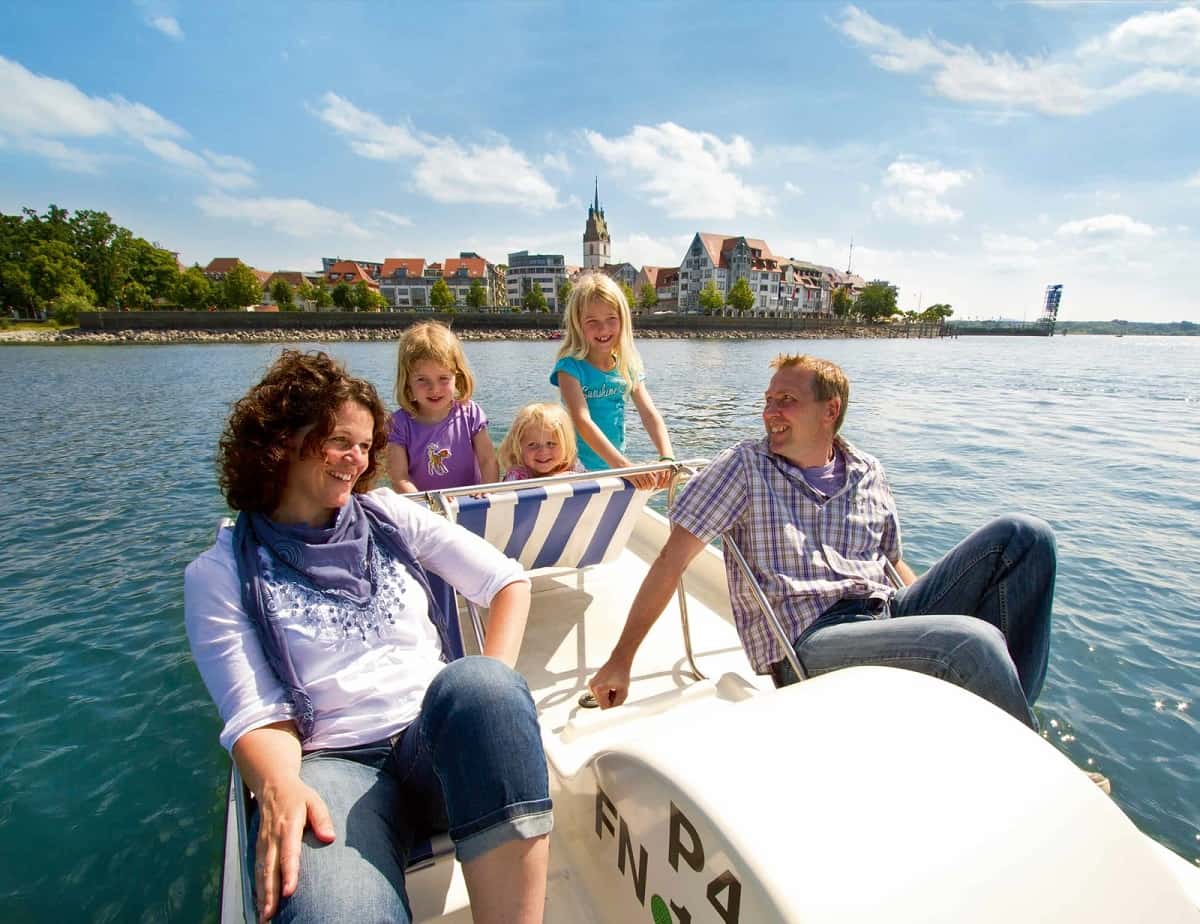 Eine Familie mit drei Mädchen fährt vor der Friedrichshafener Kulisse mit dem Tretboot auf dem Bodensee.