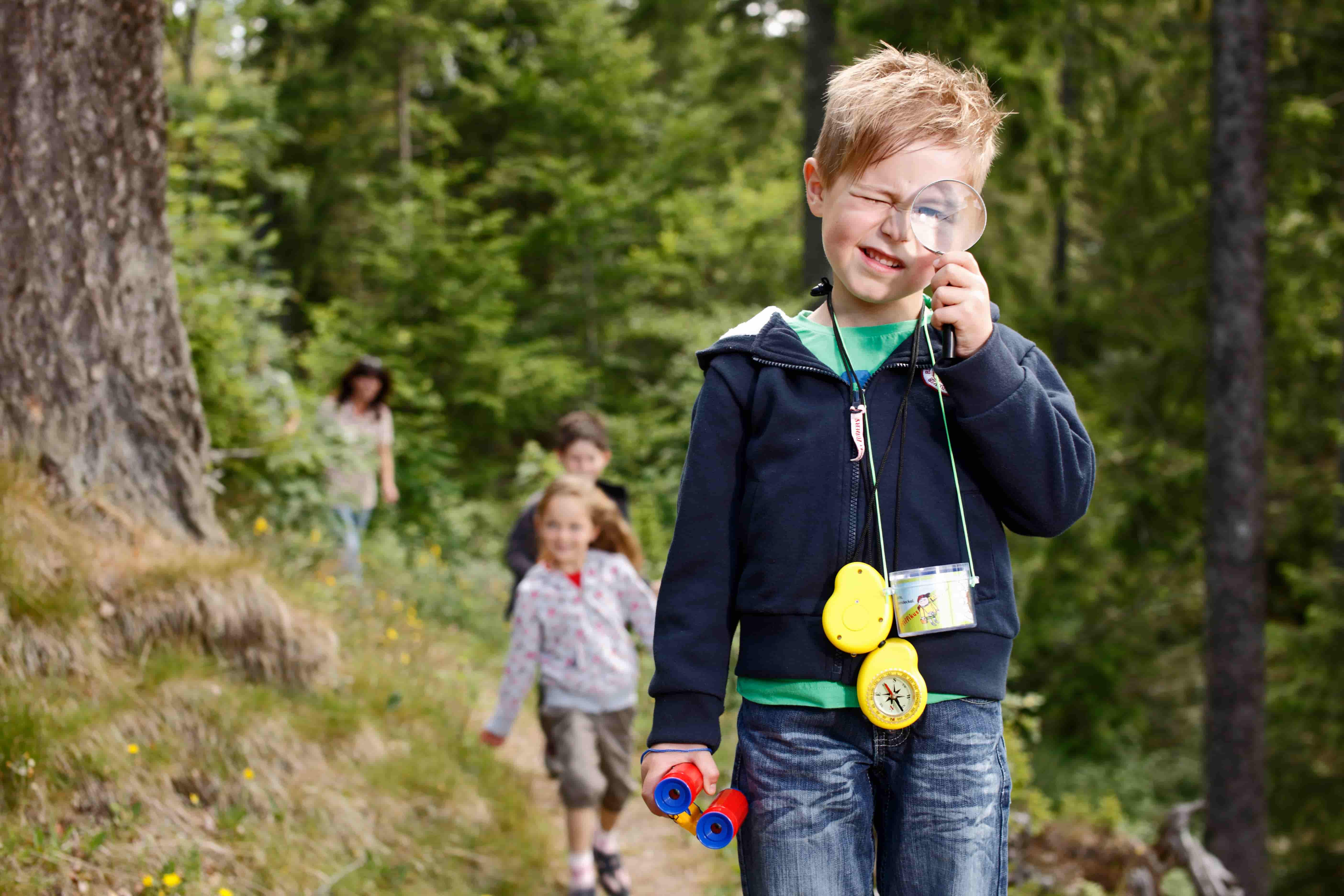 Kindern wandern auf einem Waldpfad. Das erste Kind hält ein Kinderfernglas entlang und schaut durch eine Lupe.